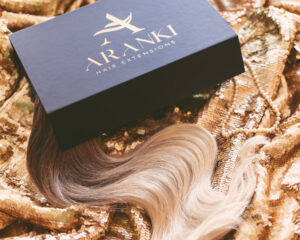 A swath of ombre clip-in extensions peeks out from a black box labeled "Aranki Hair." The box rests on a gold sequined background.
